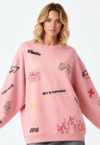 Felpa oversize rosa con vari motivi ricamati, tra cui una rosa, ali d'angelo, testo e motivi di fiamme. Texture morbida.