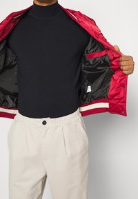 Chaqueta acolchada roja con puños de rayas crema y rojas, top negro de cuello alto debajo, y pantalones de cintura elástica de color claro.