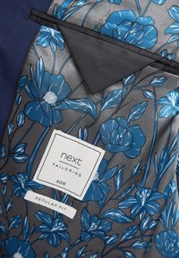 Forro de chaqueta a medida con una base gris y un estampado floral azul. Las etiquetas indican la marca y el ajuste, con un bolsillo negro contrastante.