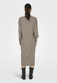Abito lungo a maglia a coste color marrone chiaro con maniche lunghe e spacchi laterali, abbinato a stivali neri alla caviglia con dettagli decorativi.
