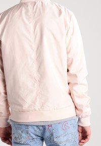 Svetle růžová bomber bunda s hladkým povrchem, žebrovanými manžetami a spodním lemem, s centrálním švem na zádech. Spárovaná se vzorovanými světle modrými džíny.