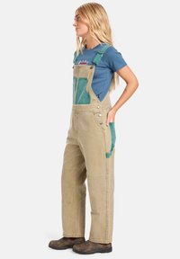 Overall color sabbia con tasche teal, bottoni laterali e spalline regolabili. Indossato sopra una camicia blu. Il tessuto sembra resistente e con una consistenza liscia.