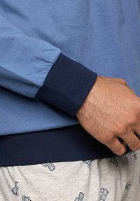 Camicia azzurra a maniche lunghe con polsini blu navy, caratterizzata da una texture liscia e un orlo aderente. Indossata sopra dei pantaloni da pigiama grigi con motivo.
