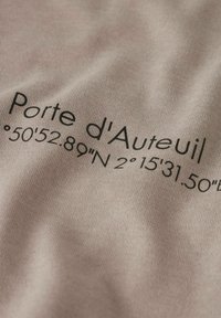 Texte sur tissu beige indiquant « Porte d'Auteuil » suivi des coordonnées géographiques 50°52,89"N 2°15'31,50"E en police noire.