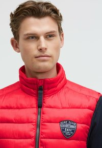 Rote gesteppte Pufferweste mit hohem Kragen, schwarzen Seitenpaneelen und einem Reißverschluss. Mit einem Logo-Patch auf der Brust.