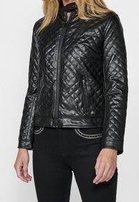 Giacca in pelle nera trapuntata con colletto alto, tasche con zip e design aderente. Presenta una texture liscia e dettagli in metallo argento.