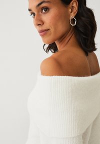 Off-the-shoulder, ribbet hvid striktrøje med blød tekstur. Har en glat halsudskæring og matchende hoop øreringe med perler.