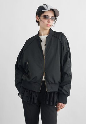 Nuori henkilö pukeutuneena mustaan vetoketjulliseen bomber-takkiin, mustaan pilkulliseen kerrospaitaan, mustiin housuihin, harmaaseen lippalakkiin ja aurinkolaseihin yksiväristä taustaa vasten.