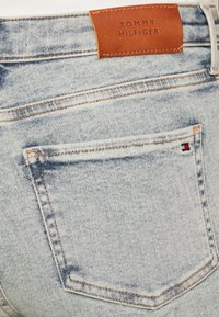 Jeans de mezclilla azul claro con un bolsillo trasero cosido y una etiqueta de cuero marrón con el logo "TOMMY HILFIGER" en relieve. Costuras en contraste evidentes.