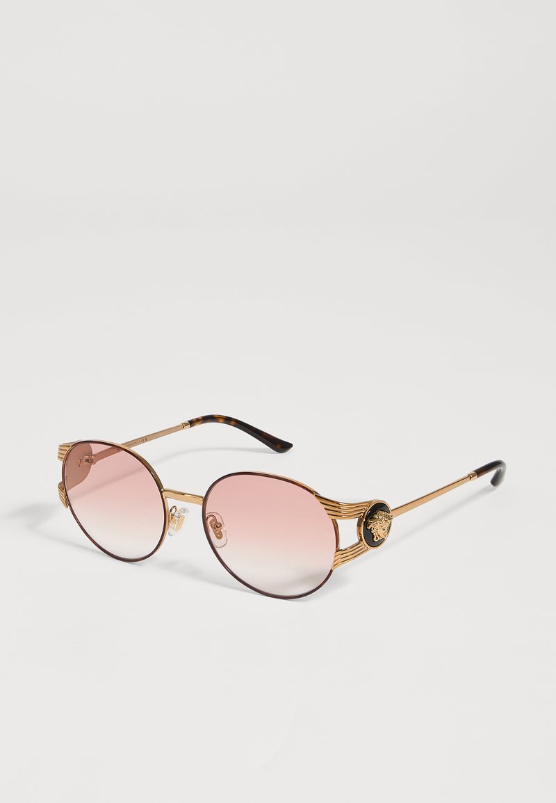 Round sunglasses with gold metal frames and pink gradient lenses, featuring a detailed emblem on each temple near the hinges.
