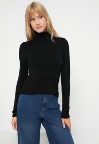 Suéter preto de gola alta em um design ajustado com mangas longas, usado com jeans de cintura alta azuis. Textura suave e detalhes mínimos.