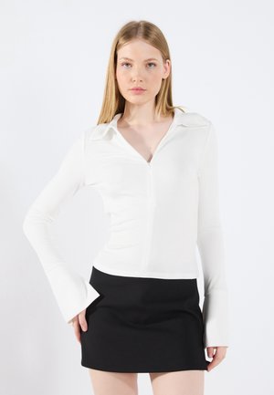 White ribbed long-sleeve top with a zip closure and wide bell sleeves, paired with a fitted black mini skirt. Smooth texture, modern design.