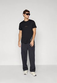 Mees, kes kannab musta graafilist t-särki, lahtisi musti pükse, valgeid tossusid ja üle suuruses musta päikeseprillipaari, seisab ühevärvilise heleda tausta ees.