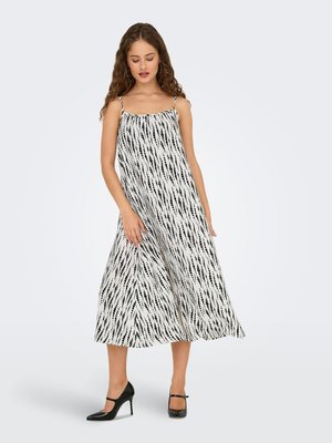 Femme portant une robe blanche longue jusqu’aux chevilles avec des rayures verticales noires à pois et des chaussures Mary Jane à talons noirs, debout sur un sol gris.