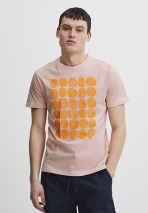 Short-sleeve pink cotton t-shirt featuring a grid of textured orange circles. Crew neck with a relaxed fit, complemented by casual dark trousers.