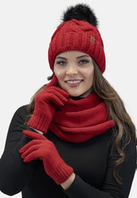 Rød strikket hue med en sort pom-pom, matchende strikket tørklæde og handsker. Har ribbet struktur og et logo-detalje på hatten.
