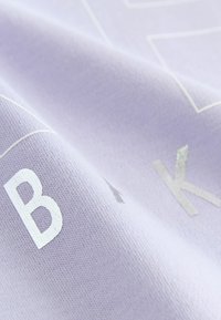 Ljust lila tyg med strukturerade vita bokstäver "B" och "K". Ytan har en mjuk, slät struktur med en lätt glans.