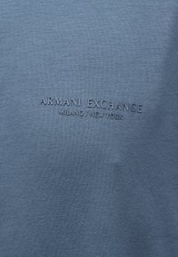 Μπλε ύφασμα με ανάγλυφο κείμενο "ARMANI EXCHANGE MILANO / NEW YORK" στο κέντρο της επιφάνειας.