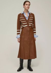 Cardigan a righe marroni sopra una camicia azzurra con colletto, abbinato a una gonna di pelle marrone con bottoni e stivali neri al ginocchio.