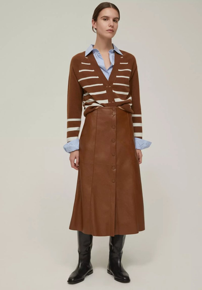 Cardigan a righe marroni sopra una camicia azzurra con colletto, abbinato a una gonna di pelle marrone con bottoni e stivali neri al ginocchio.