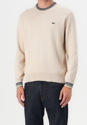 Beiger Pullover mit Rundhalsausschnitt, gestreiften, gerippten Bündchen und Kragen, mit kleinem grünen Krokodil-Logo auf der Brust, getragen zu dunkelblauen Jeans.