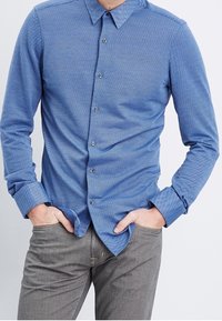 Camicia blu con bottoni, caratterizzata da un motivo a spina di pesce e colletto a punta. Maniche lunghe con polsini a bottone. Il tessuto presenta una finitura testurizzata.