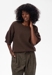 Vrouw met krullend haar in een losse bruine trui en gestructureerde bruine broek met touwtjes, poseert met één hand op haar nek en één in haar zak.