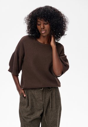 Kobieta o kręconych włosach w luźnym brązowym swetrze i teksturowanych brązowych spodniach z troczkami, pozująca z jedną ręką na szyi i drugą w kieszeni.