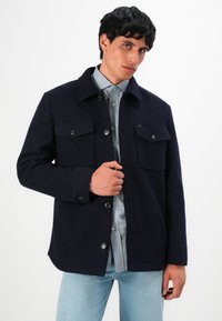 Mann mit dunklem Haar, der eine navyfarbene, geknöpfte Jacke über einem hellblauen Hemd und hellblauen Jeans trägt und mit einer Hand die Jacke zurechtrückt.
