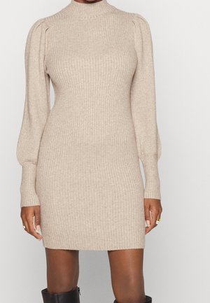 Beige ribstrikket sweaterkjole med lange pufærmer og høj hals, båret med sorte kniehøje støvler.