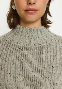 Pulover tricotat de culoare gri deschis, cu un guler înalt dintr-un material striat, având un model texturat și stropi de culoare mai închisă pe întreaga țesătură.