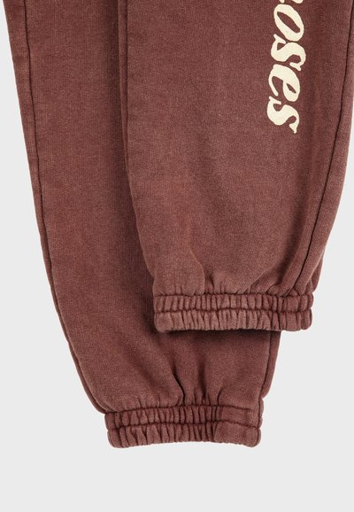 Pantalones deportivos marrones con puños elásticos y texto de color crema parcialmente visible en la pierna izquierda, sobre un fondo neutro.