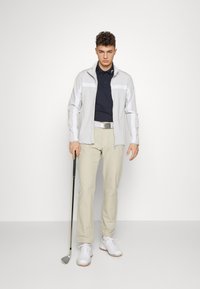 Chaqueta gris claro con cremallera y detalles en blanco, camiseta de polo azul marino, pantalones beige y zapatos de golf blancos. Sosteniendo un palo de golf, de pie contra un fondo blanco.