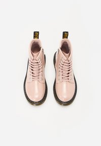 Botas de tobillo de charol rosa con suela negra, ojales plateados y cordones; cremallera lateral y etiqueta amarilla en el talón. Textura suave y brillante.