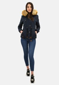 Vrouw in een marineblauwe winterjas met een bontkraag, blauwe skinny jeans en zwart-witte sneakers, staande met haar handen in haar zakken.