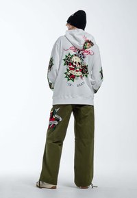 Sudadera gris con gráficos de cráneo y rosa, combinada con pantalones verde oliva que presentan motivos similares. Zapatillas blancas completan el look.