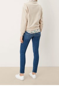 Sweat-shirt beige avec un col montant, grand panneau arrière, chemise à fleurs en dessous, jean skinny bleu et baskets blanches.