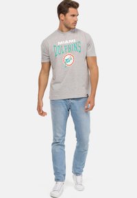 Šedé bavlněné tričko s nápisem "Miami Dolphins" v tyrkysové a bílé barvě, s grafikou delfína uvnitř kruhové pečeti. V kombinaci se světle modrými džíny.