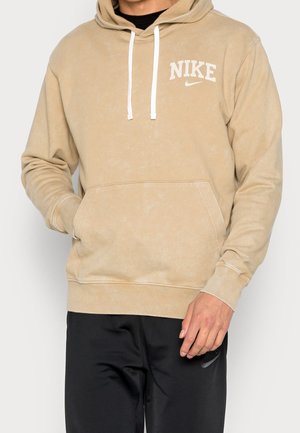 Beige Nike-hoodie met witte trekkoorden en logo op de borst, gedragen met zwarte Nike-broek. Handen deels zichtbaar.