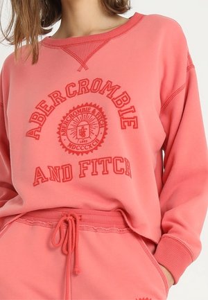 Žena nosí korálově růžovou mikinu Abercrombie and Fitch a odpovídající tepláky s tahem v pase, ruka v kapse.