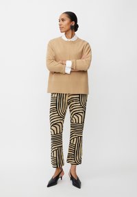 Beiger Strickpullover mit strukturierter Oberfläche, kombiniert mit gemusterten beige- und schwarzfarbenen Hosen mit geschwungenen und gestreiften Designs. Schwarze spitze Schuhe.
