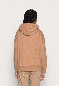 Sudadera con capucha de tela marrón claro, con un corte relajado, hombros caídos y dobladillo acanalado. Textura suave sin patrones visibles.