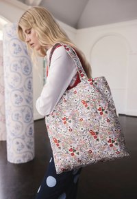 Plavolaska ženska z belo srajco in rdečo jopico nosi veliko torbo z vzorcem cvetlic in simbolov kart na rami v svetlem notranjem prostoru.