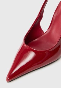 Pompe à talon aiguille en cuir verni rouge avec un bout pointu et un design découpé sur les côtés. Fini lisse et brillant avec une semelle noire.