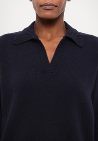 Personne à la peau foncée portant un pull en laine bleu marine avec col et encolure en V sur fond clair uni.