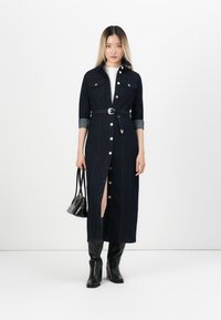 Robe chemise en denim, bleu foncé, manches longues avec poignets, fermeture à boutons sur le devant, taille ceinturée, fente sur le côté, associée à des bottes noires hauteur genou et un petit sac.