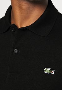 Svart pikétröja med en ribbad textur, utrustad med en tvåknappsknäppning och en liten broderad grön och vit krokodil-logotyp på vänster bröst.