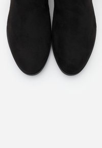 Botines de ante negro con textura suave y diseño de puntera redonda, que presentan una suela baja y plana sin herrajes o acentos visibles.