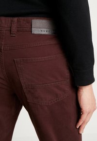 Pantaloni in tessuto bordeaux con texture liscia, tasca posteriore e una toppa in pelle scura con marchio impresso sulla cintura.