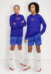 Modré mikiny s kapucí a oranžovým logem "CR7" spolu se vzorovanými modrými šortkami. Děti mají bílé ponožky Nike a fotbalové boty s červenými akcenty.